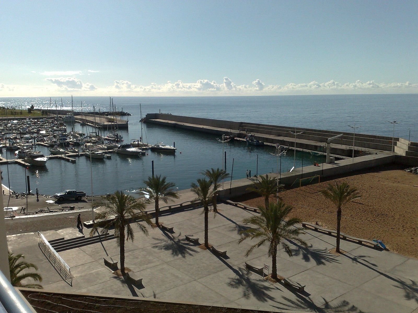 Calheta marina from above, yachts moored inside two concrete breakwaters with a fishing boat alongside the outer arm, palms and a paved promenade in front, and pale golden sand at the right of the frame.