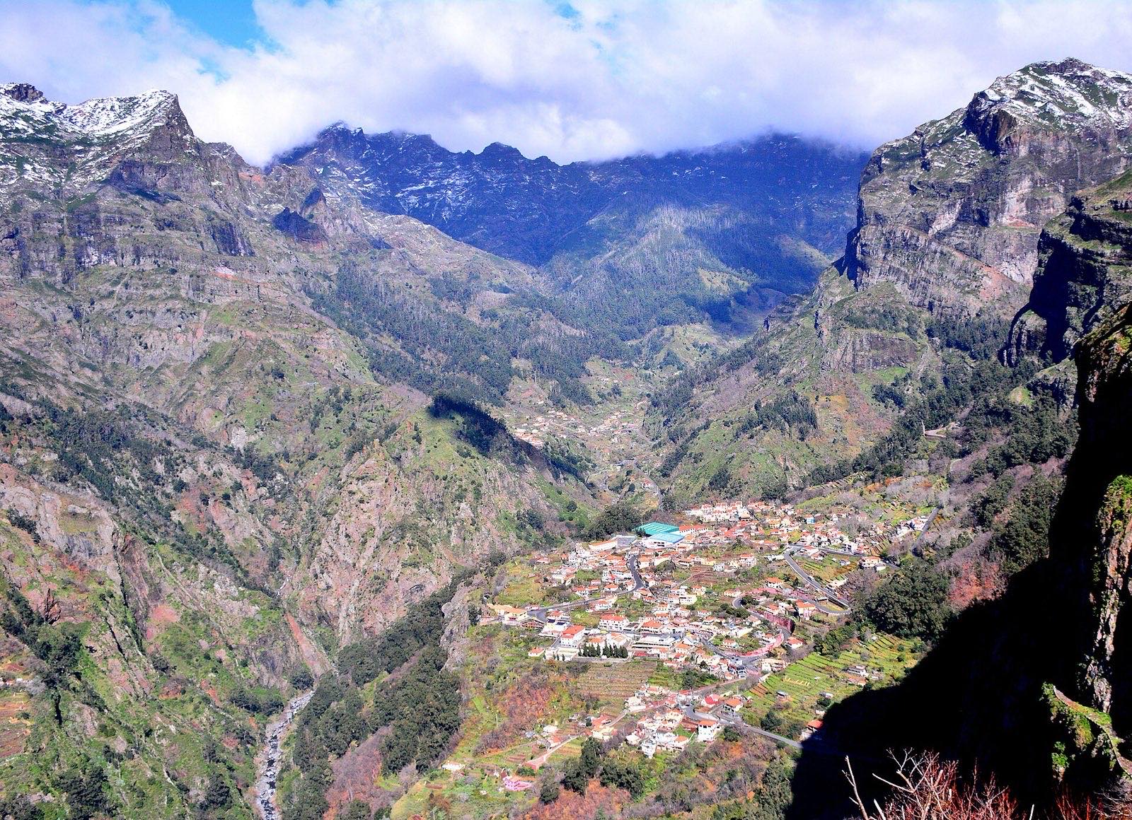 Curral das Freiras seen from the rim high above: white orange-roofed houses on the valley floor, sheer green and red rock walls closing in on both sides, and snow lying on the peaks at the head of the valley.