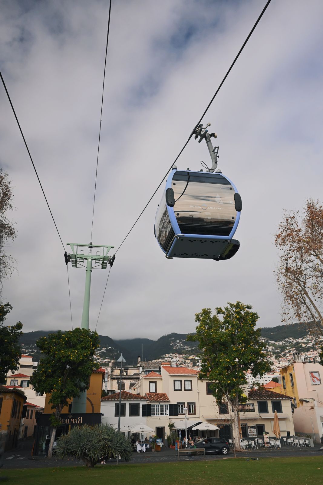 A cable car cabin hanging from its wire above Funchal’s Old Town, tiled roofs and café awnings below it, houses climbing the hillside behind and cloud lying along the ridge above the city.
