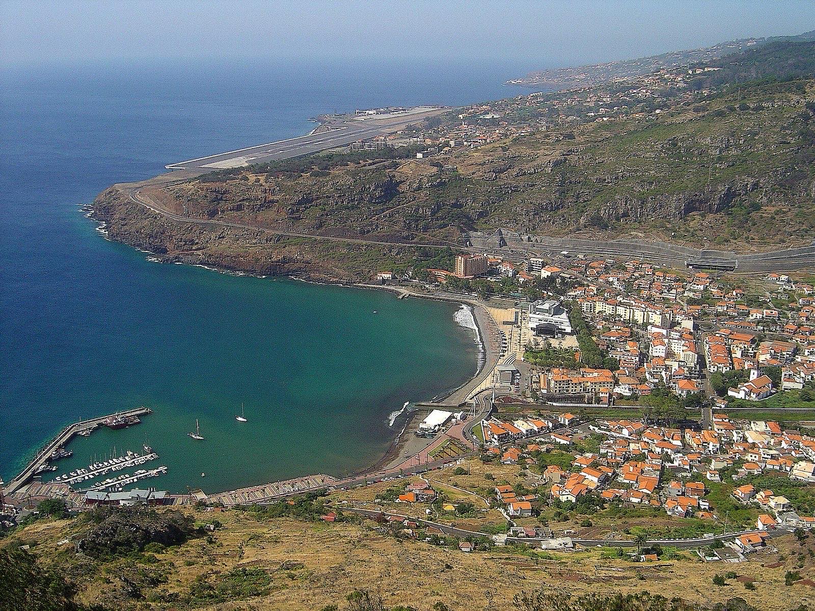 Machico from the hill above the bay: a curve of pale sand and a marina behind a breakwater, the town packed along the shore, and the airport runway on its embankment further along the coast.