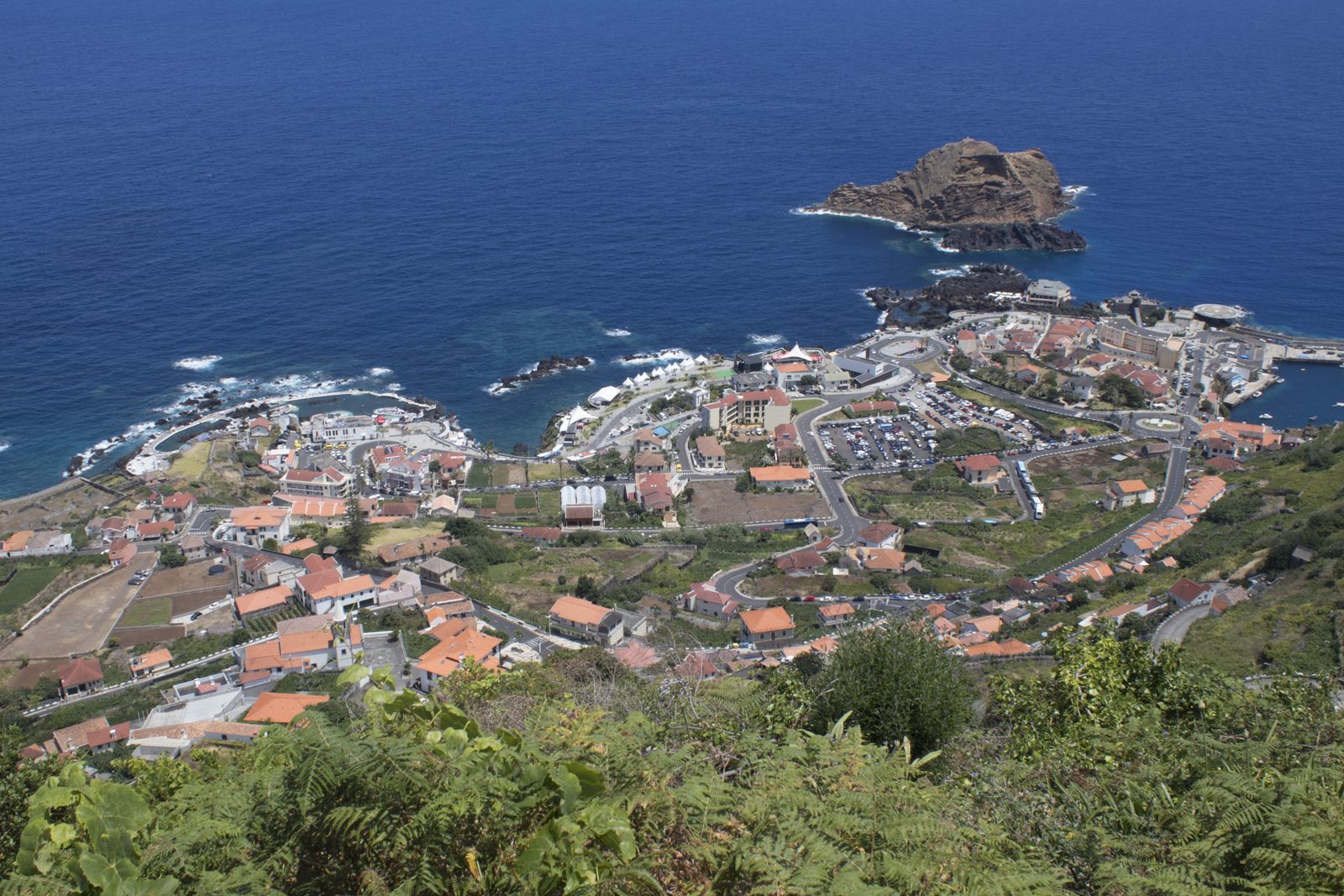 Porto Moniz from the hillside: the village spread over a low headland, bathing pools cut into black rock at the water line, white surf breaking along the shore and a flat-topped islet standing offshore.