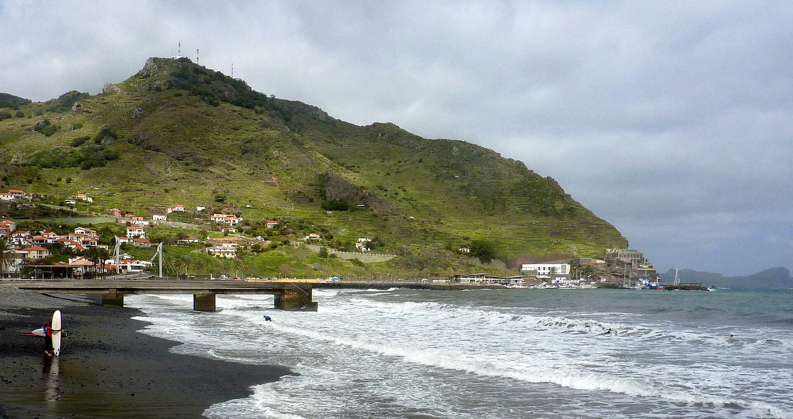 Ein dunkler Kieselstrand bei Machico unter schwerer Bewölkung, Wellen brechen auf ganzer Länge, ein niedriger Betonsteg auf Stützen, ein Surfer mit Brett am Wasser und dahinter der grüne Berg Pico do Facho über dem Ort.