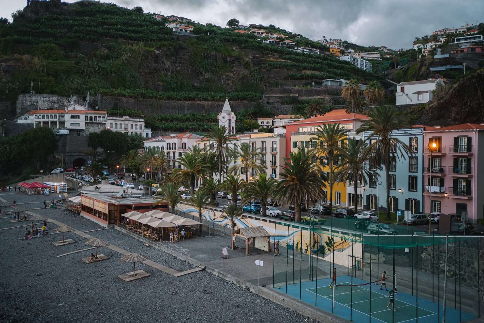 Strohschirme und Liegen auf grauem Kies in Ponta do Sol in der Dämmerung, Restaurantterrassen und ein beleuchteter Ballplatz entlang der Front, pastellfarbene Häuser und ein Kirchturm unter Bananenterrassen.