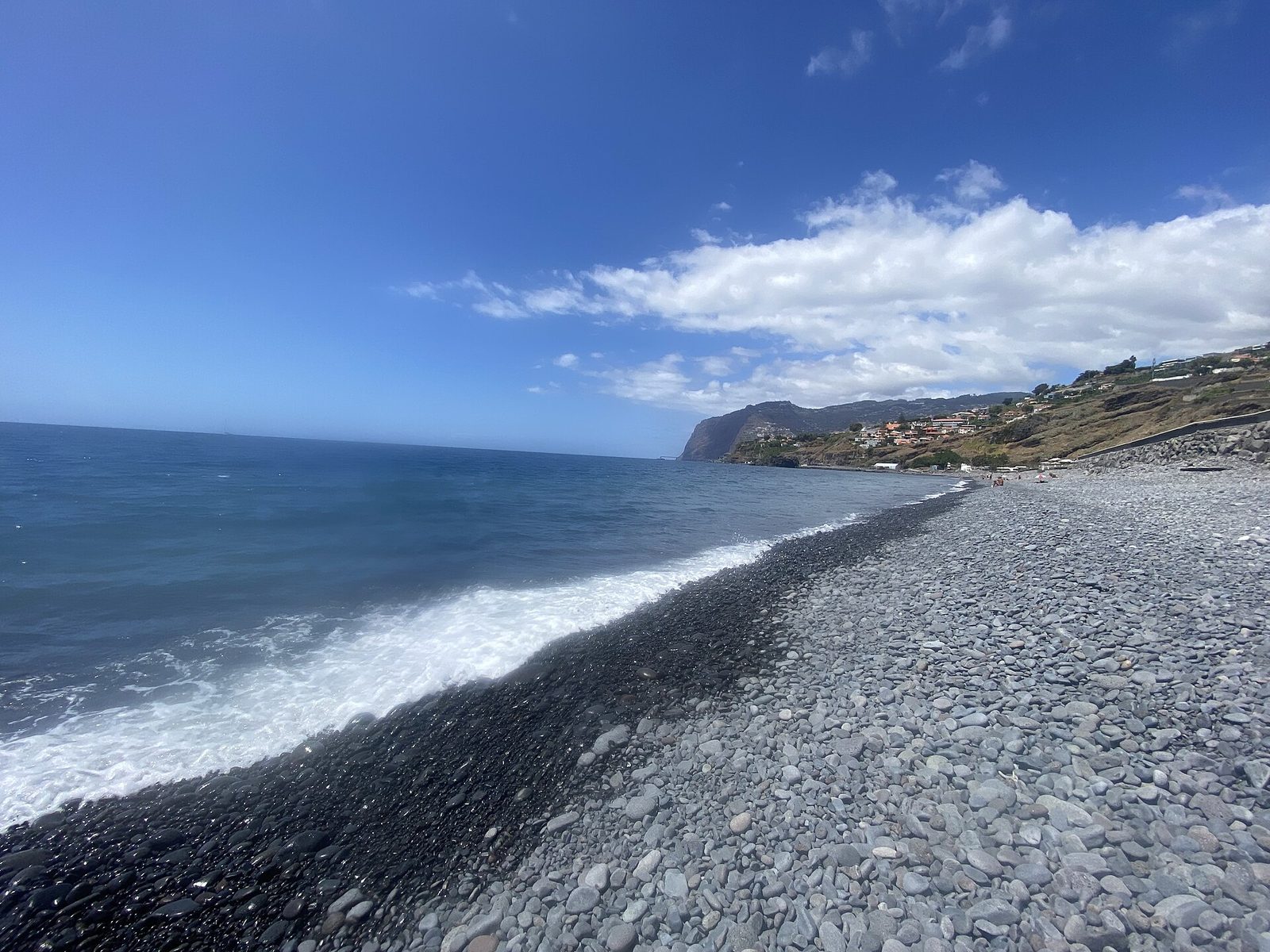 Ein langer Strand aus grauen, rundgeschliffenen Kieseln, der zu einem Streifen nassen schwarzen Sandes an der Brandungslinie abfällt, links glattes blaues Meer und in der Ferne ein Kap mit Häusern am Hang.