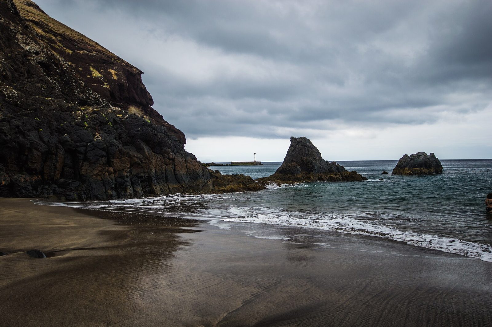 Ein Bogen dunklen braungrauen Sandes bei Prainha unter Gewitterwolken, links ein rotbraunes Vulkankap, Lavafelsen im flachen Wasser und in der Ferne ein Hafenleuchtfeuer.