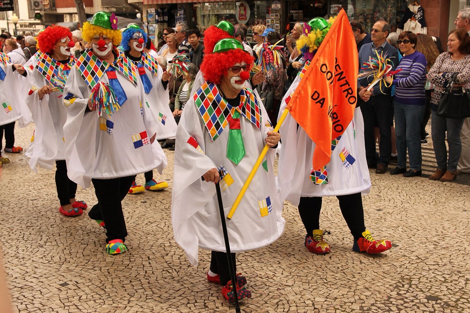 A group of carnival walkers in white harlequin capes, red curly wigs and clown make-up crossing Funchal’s black-and-white cobbles in daylight, one carrying an orange banner, with spectators lining the shopfronts behind them.