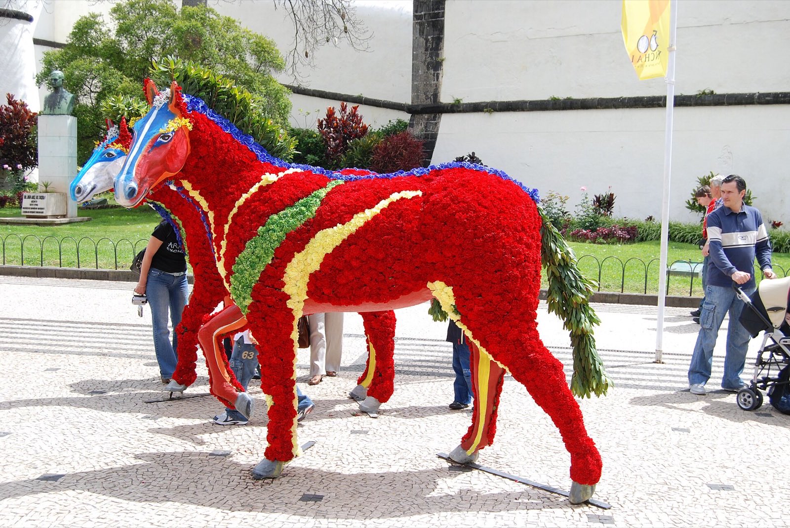 Two life-size horse sculptures covered entirely in red, yellow, green and blue flowers, standing on a Funchal pavement beside a whitewashed church wall, with people walking past them.