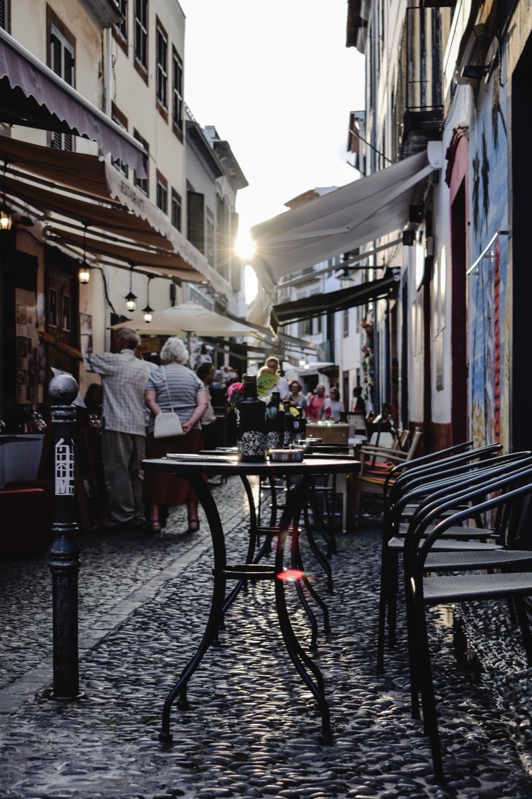 Une ruelle pavée étroite de la vieille ville de Funchal au soleil rasant, tables de restaurant installées sur le sol, auvents au-dessus et panneaux peints sur le mur.