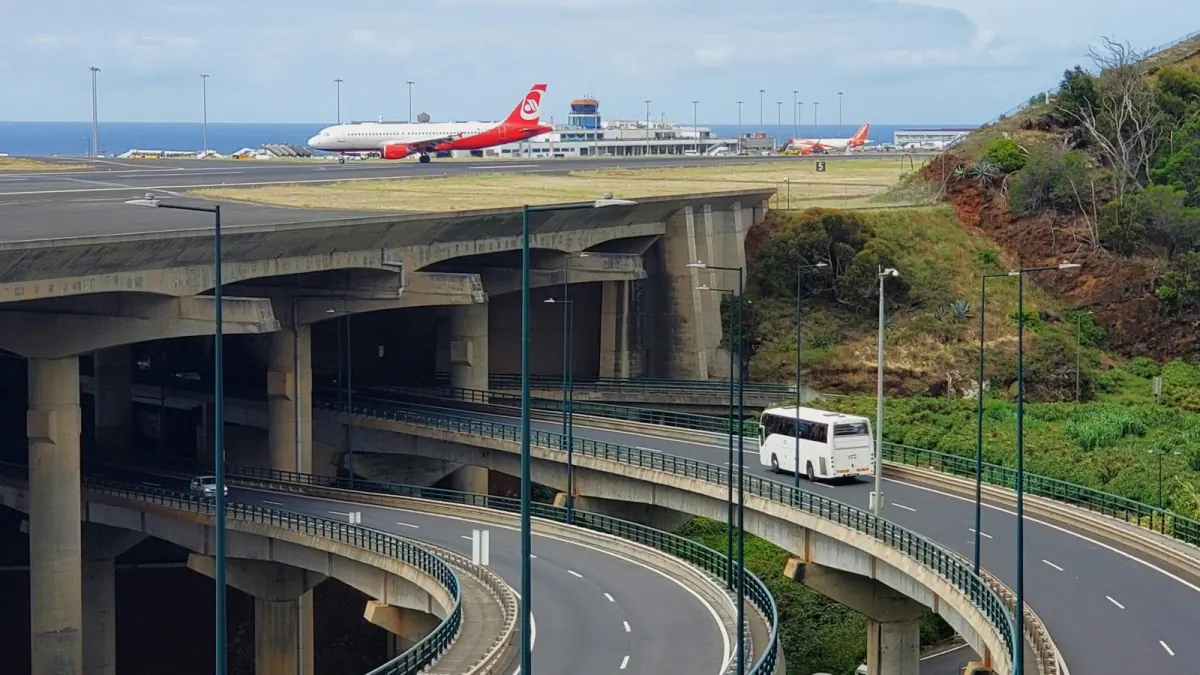 Two airliners beside Madeira Airport’s control tower, with the runway deck and its concrete supports above the looping coast-road interchange.