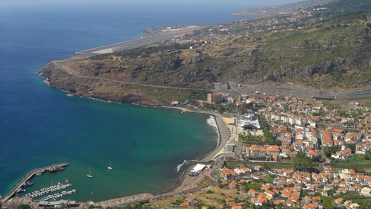 A wide view over Machico bay, with the marina and pale beach below the town and Madeira Airport’s runway stretching along the coast beyond.