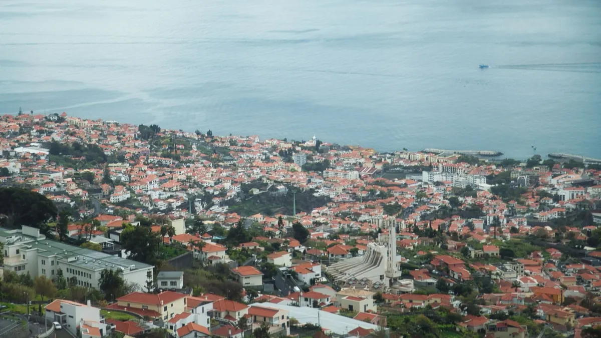 A wide view down across Funchal’s dense orange-tiled neighbourhoods to the harbour and flat grey Atlantic, with steep roads threading between the buildings.