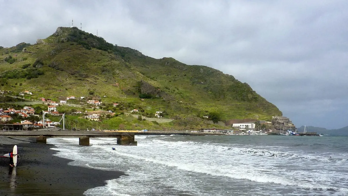 Heavy cloud over Machico’s green Pico do Facho, with rough waves crossing the dark shingle shore and a surfer carrying a board beside the low concrete pier.