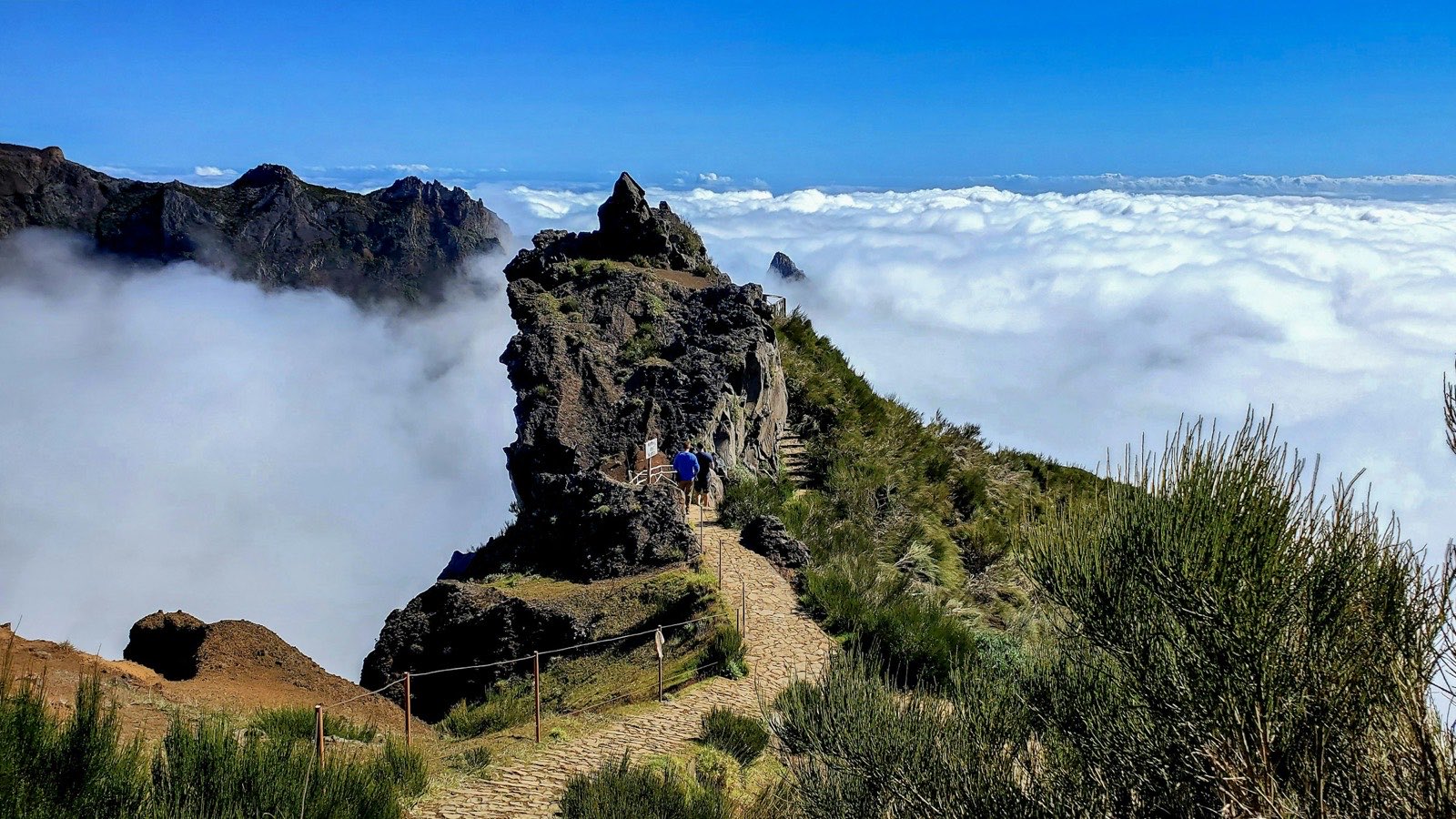 Two walkers on the paved ridge path from Pico do Arieiro, the trail running along a knife-edge of dark rock with a solid white sea of cloud filling the valleys on both sides.