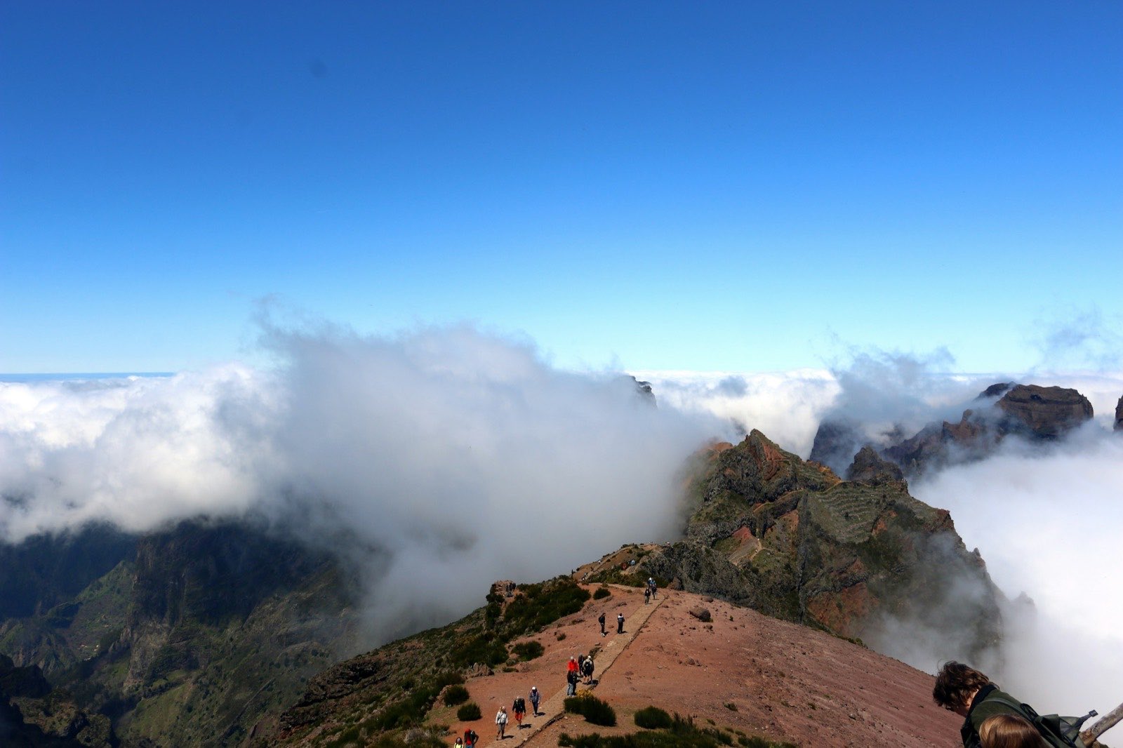 Walkers strung out along a red earth ridge below Pico do Arieiro as a bank of cloud spills over from the left, the peaks standing clear above it.
