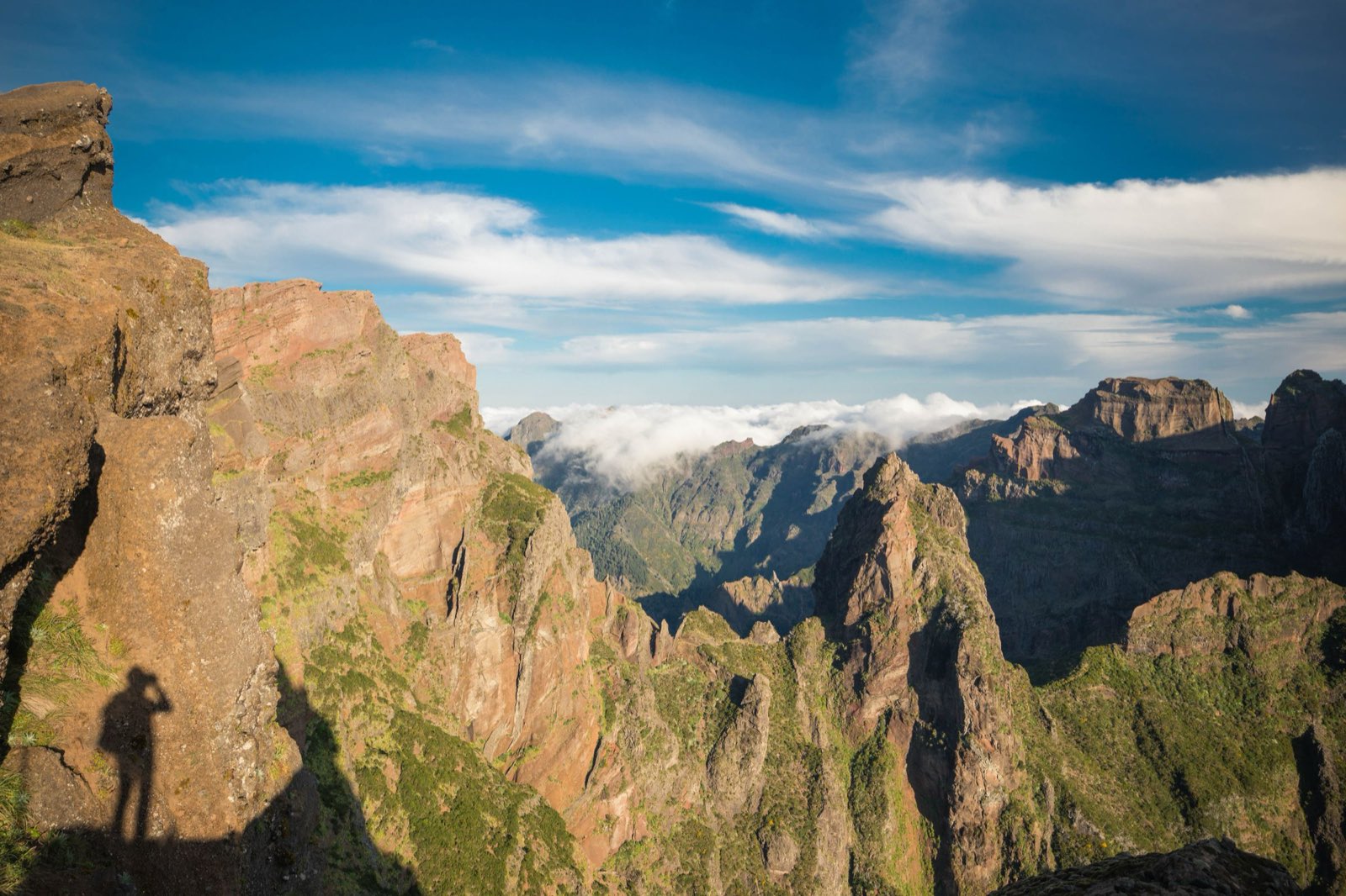 The view from Pico do Arieiro across red and green volcanic spires and ravines, with a low band of cloud lying in the middle distance.