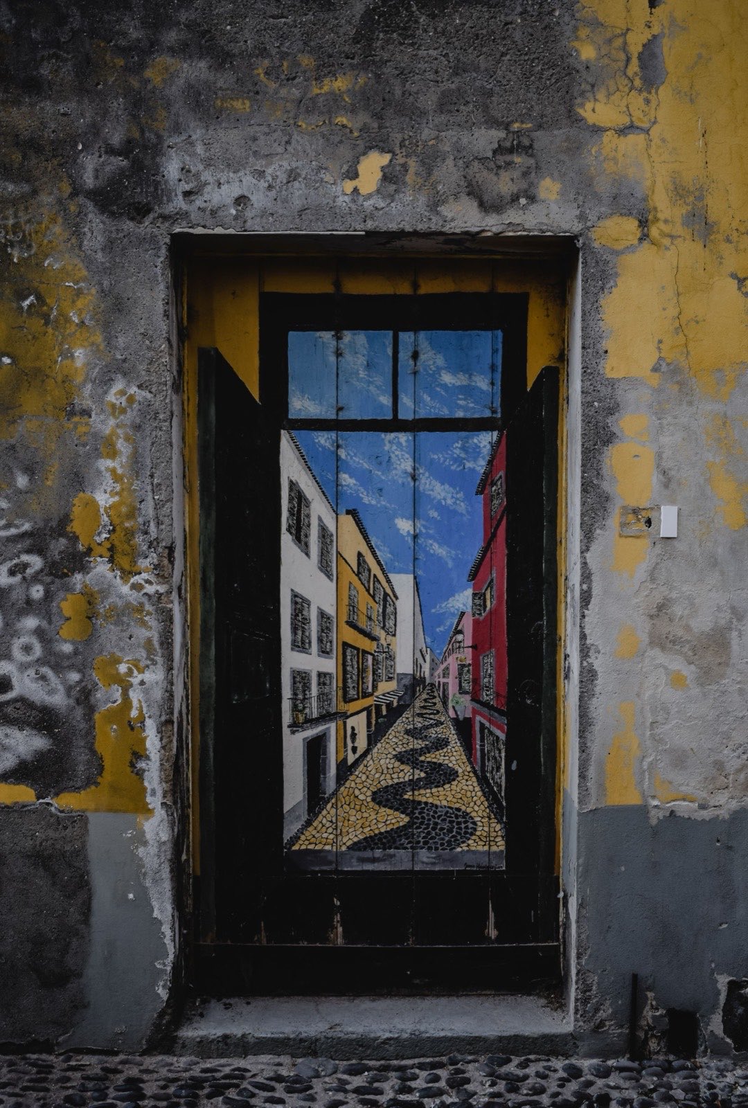 Eine verwitterte Holztür, bemalt mit dem Blick in eine madeirische Gasse — weiße, gelbe und rote Häuser über schwarz-weißem Pflaster — in einer abblätternden gelben Wand.
