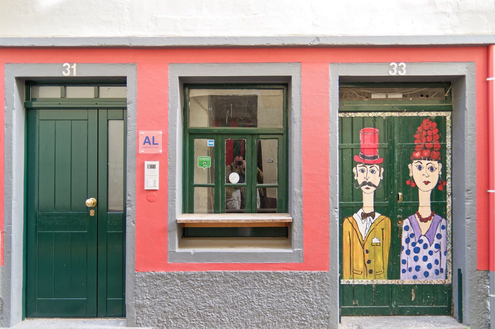 Zwei Türen an einer korallenrosa Fassade in Funchal: eine schlicht dunkelgrün, die nächste bemalt mit einem Paar mit Zylinder und rotem Kopfschmuck.