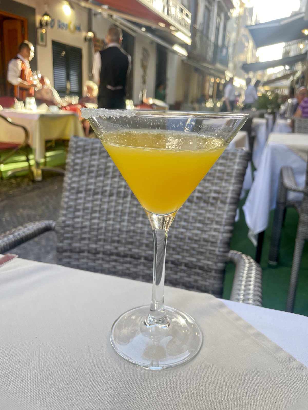 Un verre à pied givré de sucre rempli de poncha orangée, sur une nappe blanche à une terrasse de Funchal, avec des serveurs et des stores éclairés derrière.