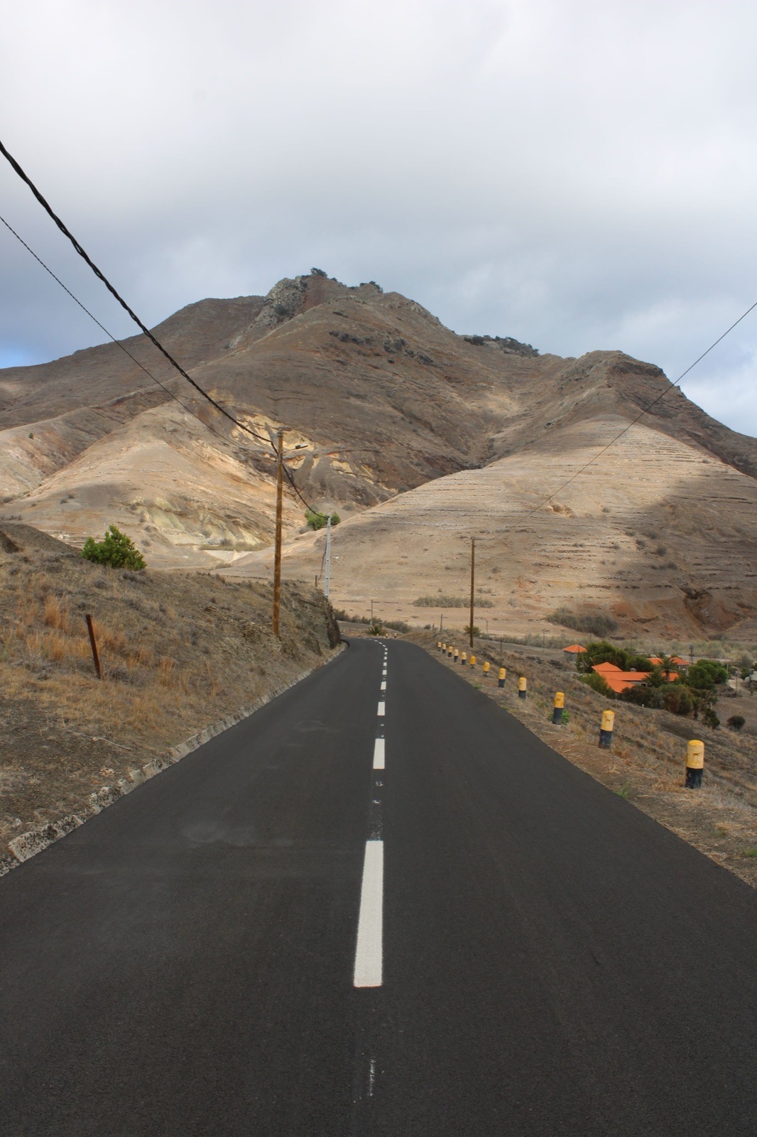 Eine leere zweispurige Straße verläuft schnurgerade auf kahle, staubfarbene Hügel auf Porto Santo zu, mit Freileitungen, einigen Pinien und orangeroten Dächern am Fuß des Hangs.