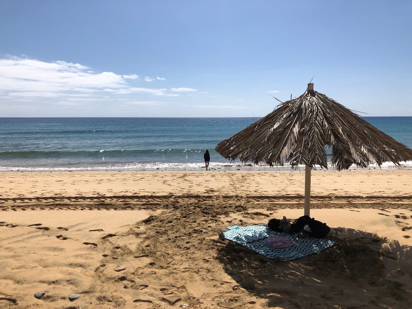 Ein Sonnenschirm aus Palmwedeln und ein Handtuch auf dem weiten, leeren goldenen Sand von Porto Santo, eine Person am Wassersaum und dahinter der blaue Atlantik.