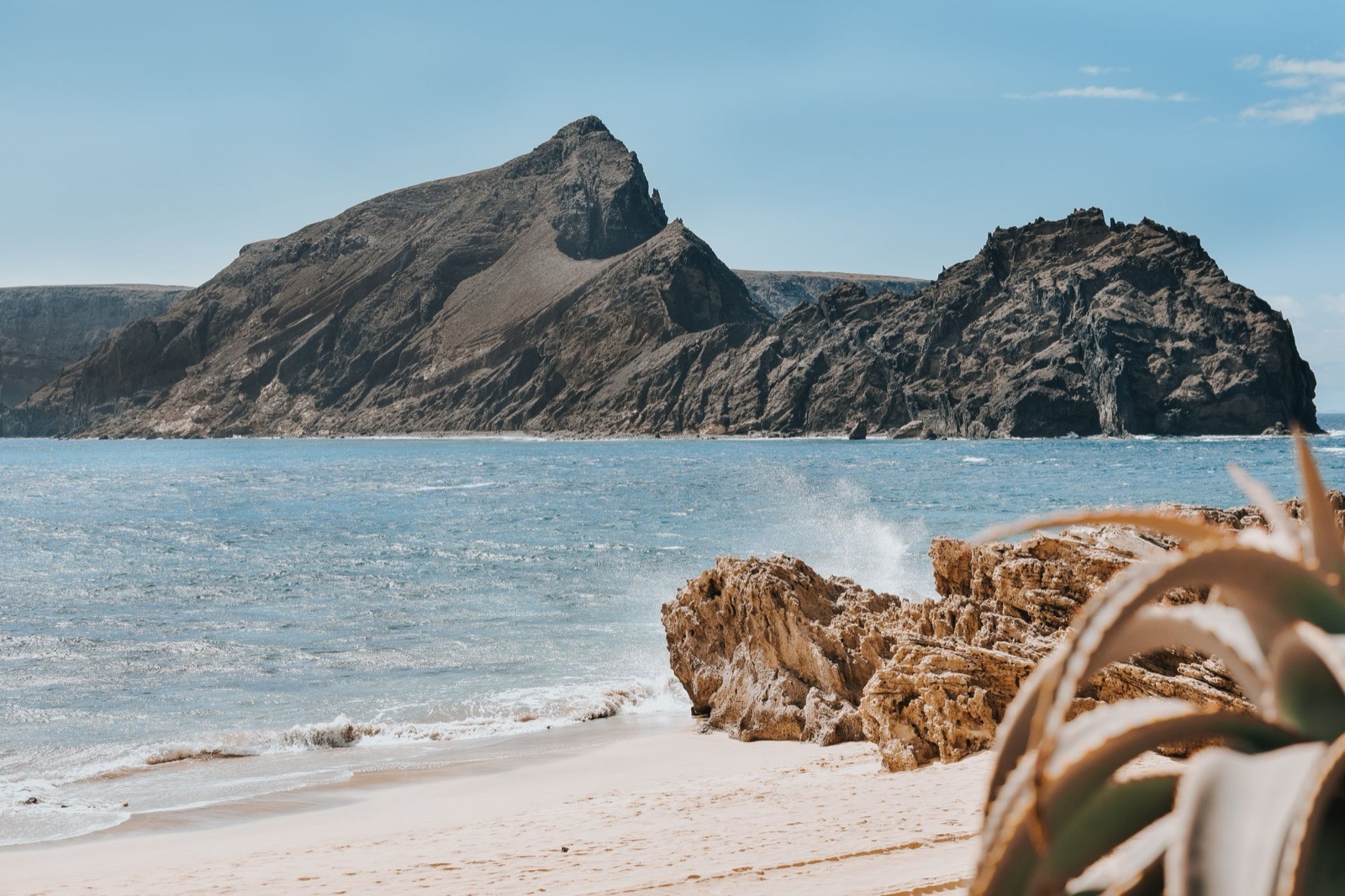 Das südliche Ende des Sandstrands von Porto Santo: Brandung läuft an einem honigfarbenen Felsen vorbei den Strand hinauf, dahinter ein hohes braunes Kap und ein vorgelagerter Felsen.