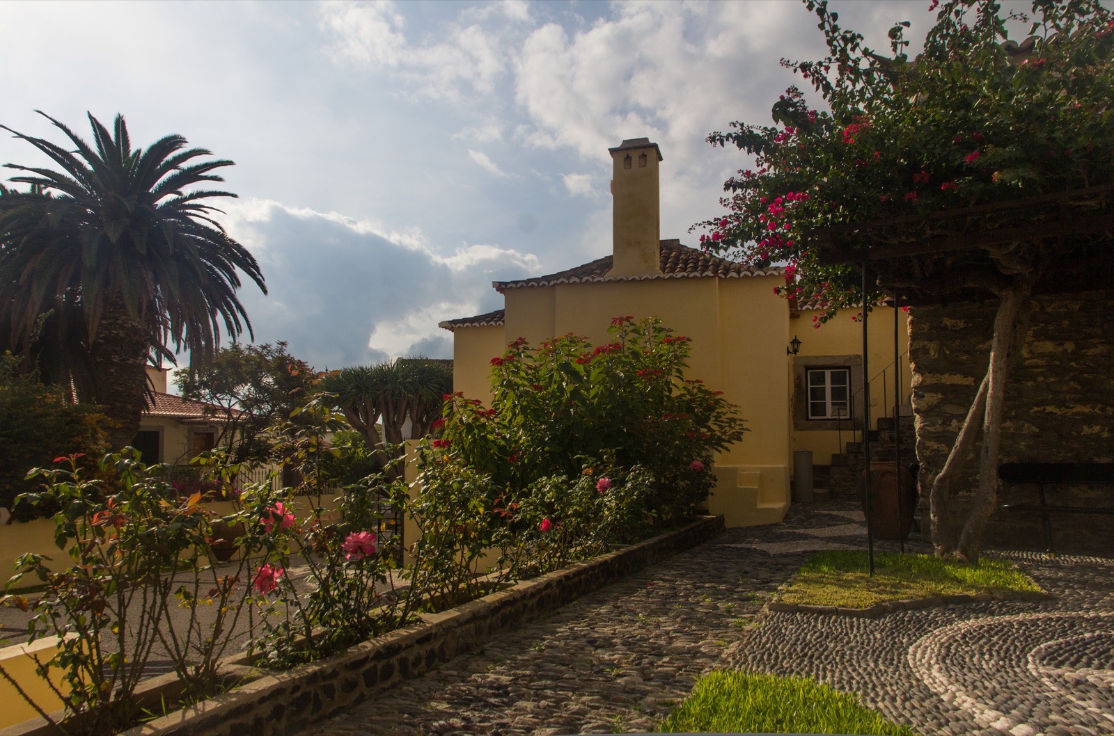 Der gepflasterte Gartenweg der Casa Colombo in Vila Baleira führt an Rosen und Bougainvillea vorbei zu einem blassgelben Haus mit Ziegeldach und hohem weißem Schornstein.