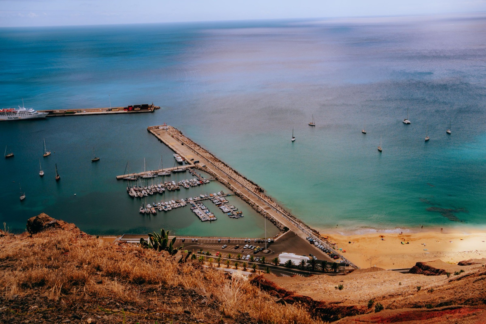 Der Hafen von Porto Santo von oben: eine weiße Fähre mit dem Schriftzug Porto Santo Line an der äußeren Mole, innerhalb des Wellenbrechers ein Yachthafen, rechts beginnt der Strand.