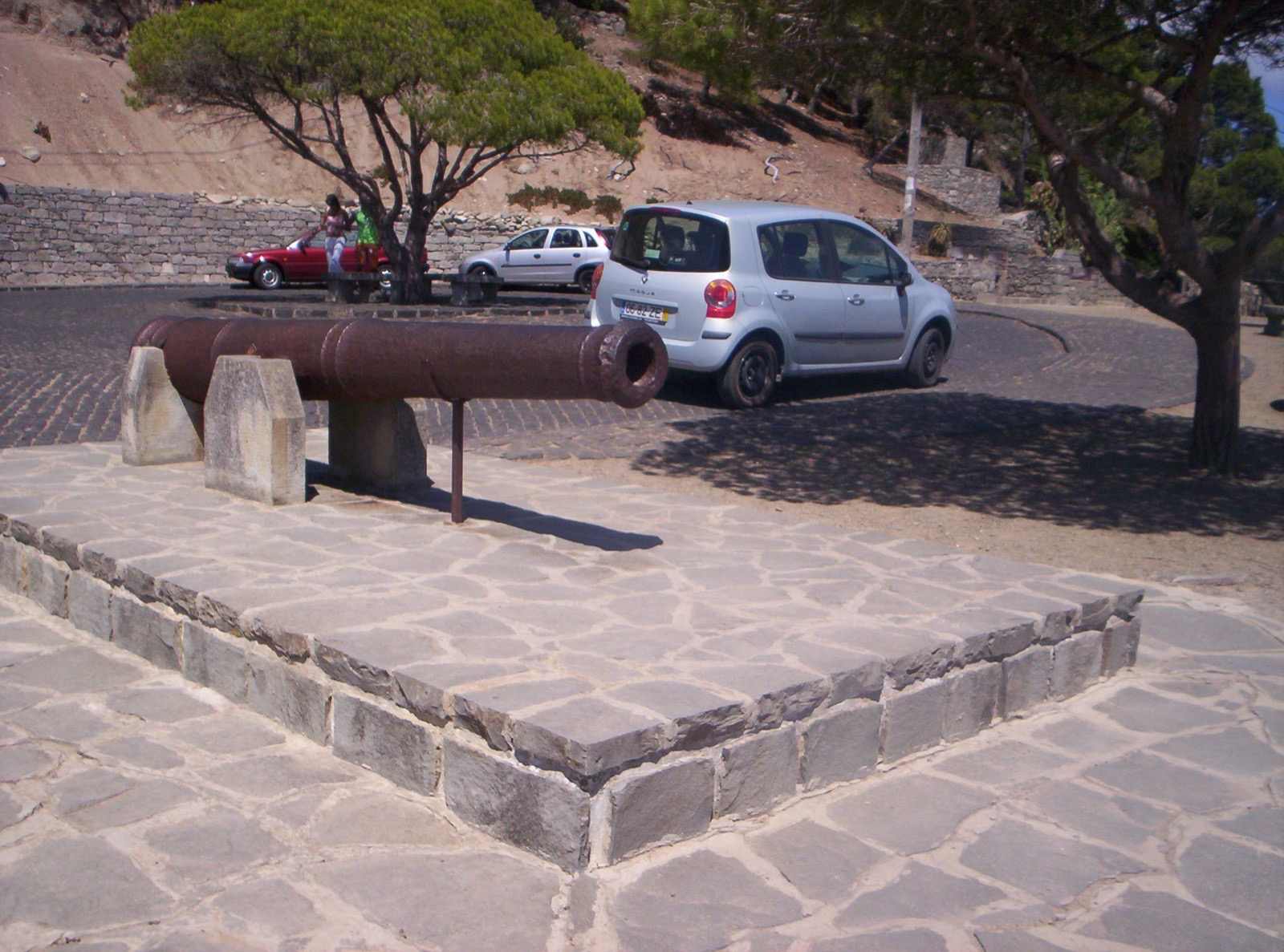 Eine verrostete eiserne Kanone auf einem Steinsockel am Aussichtspunkt Pico do Castelo, dahinter parkende Autos, Schirmpinien und der trockene Hang.