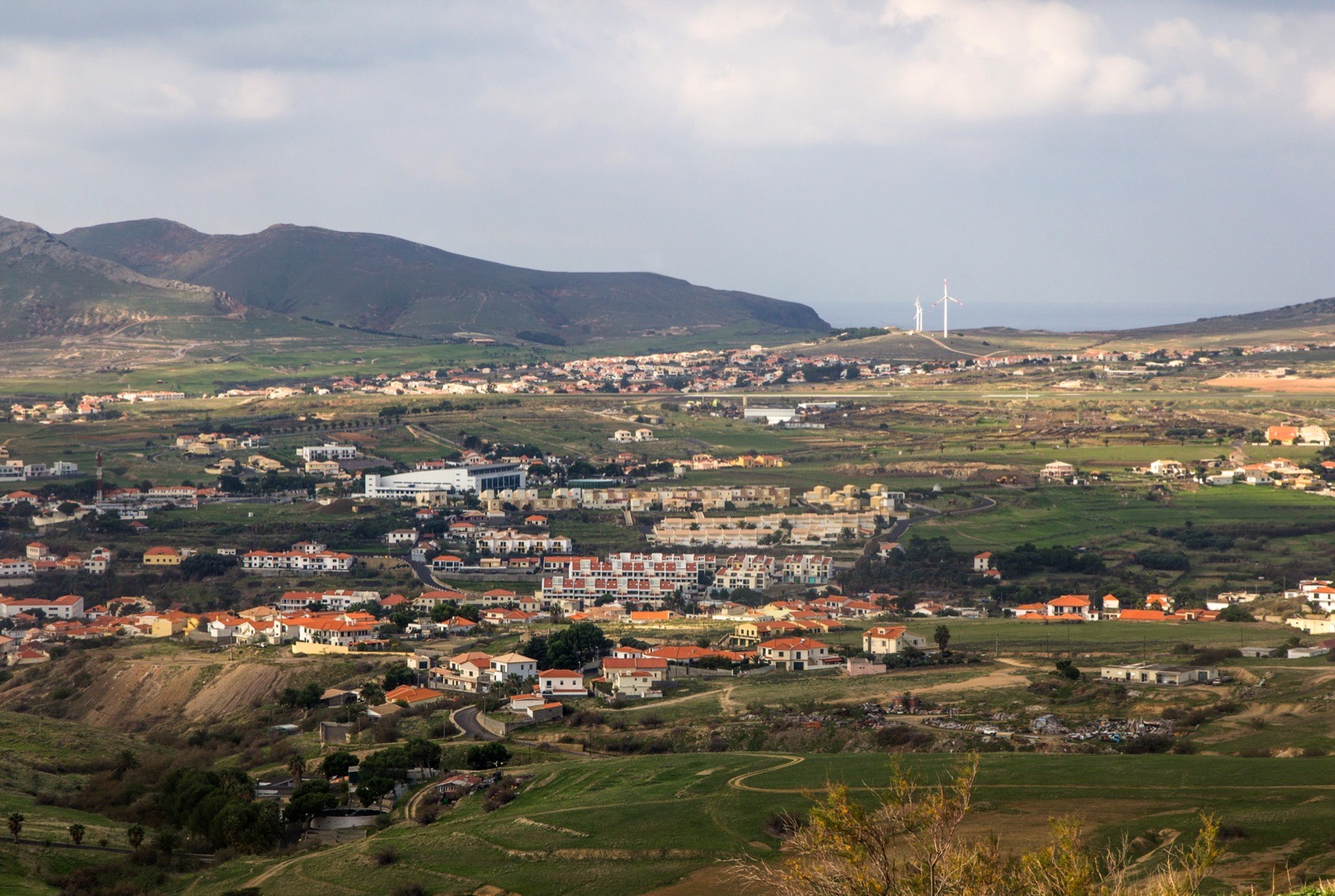 Blick von oben über Vila Baleira: weiße Häuser mit orangen Dächern auf einer grünen Ebene, dahinter die Start- und Landebahn und zwei Windräder auf dem Kamm, darüber kahle Hügel.