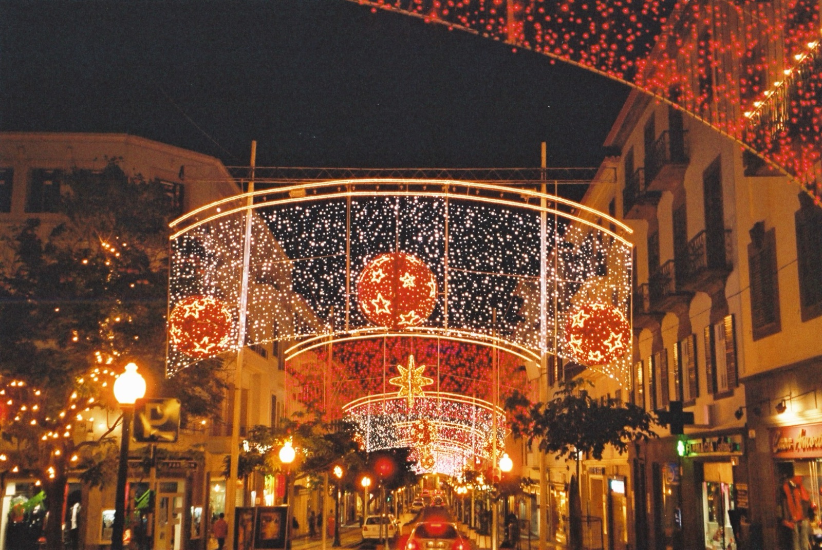 Funchal la nuit sous des guirlandes de Noël, des figures lumineuses au-dessus d’une rue animée et des vitrines éclairées derrière la foule.